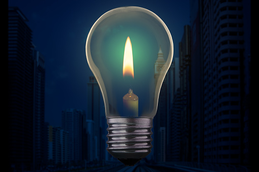 Light bulb with candle burning inside against a modern city district without electricity at night. Emergency blackout concept.