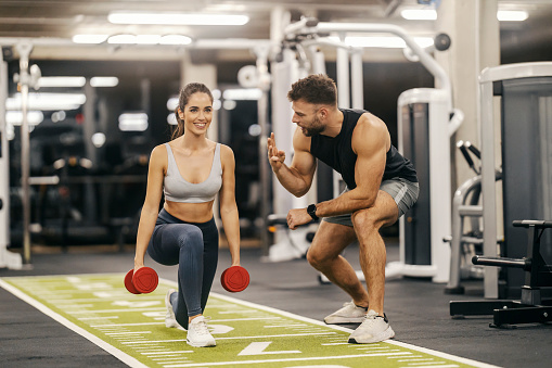 A sportswoman in shape is doing lunges with dumbbells while her trainer is counting in a gym.