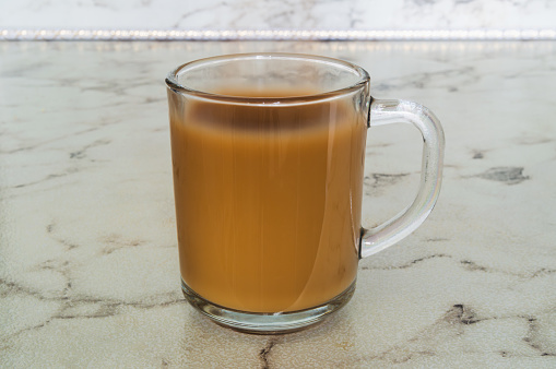 A glass cup of coffee sits on a marble countertop. The coffee is brown and he is freshly brewed. The cup is filled to the brim, and the liquid inside is swirling around the edges