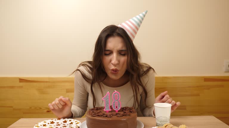 The girl is sitting in front of a table with a festive cake, in which a candle in the form of the number 18 is burning, which she blows out. Birthday celebration concept.