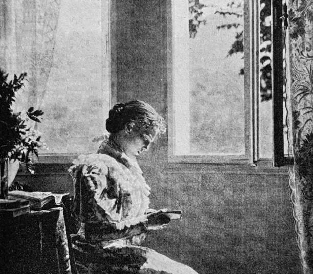 istock Young woman sitting in living room at an open window and reading a book 2111884434