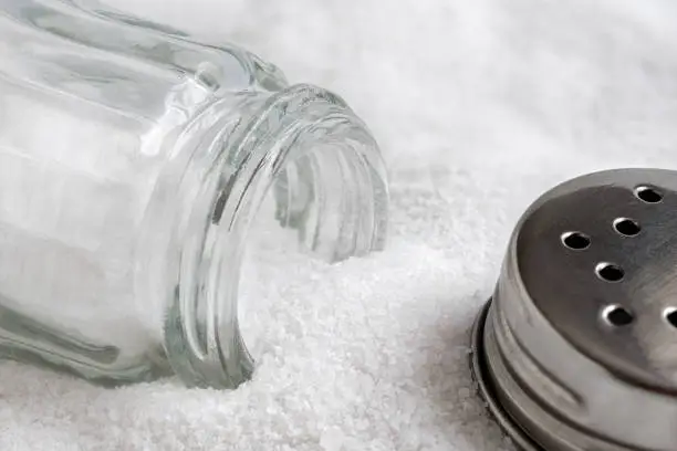 Glass salt shaker and metal lid lying on top of sea salt.