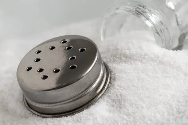 Glass salt shaker and metal lid lying on top of sea salt.