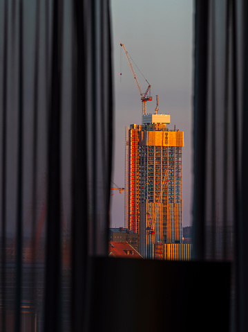 The warm glow of sunrise bathes a skyscraper under construction in Gothenburg, Sweden. Viewed from an interior perspective, the silhouette of curtains frames the emerging edifice, highlighted by the suns first light.