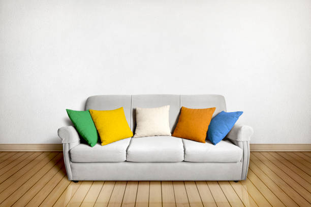 Interior background of a living room with a bright gray three-seater sofa with armrests and five colored cushions in front of a textured white wall and on a light wooden floor.