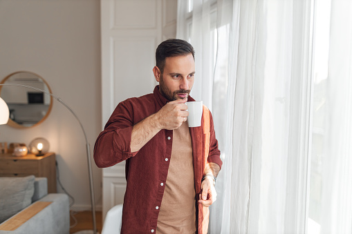A relaxed young adult man having a cup of in the morning while looking through a window and relaxing at home.