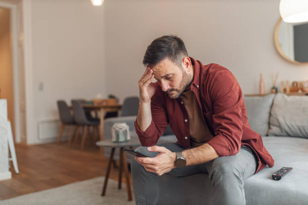 An anxious adult man reading bad news on his phone while sitting on a couch A worried adult man reading bad news and text messages on his phone while sitting on a couch in his apartment. edgy man stock pictures, royalty-free photos & images