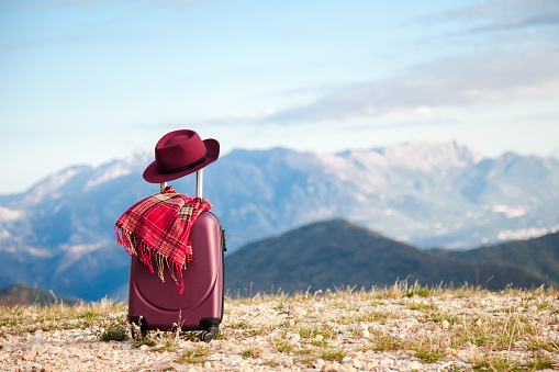 Suitcase with felt burgundy hat and plaid scarf in mountains. Concept of travel, autumn vacation, female tourism, trip, adventure. Nature background of amazing landscape with blue sky.