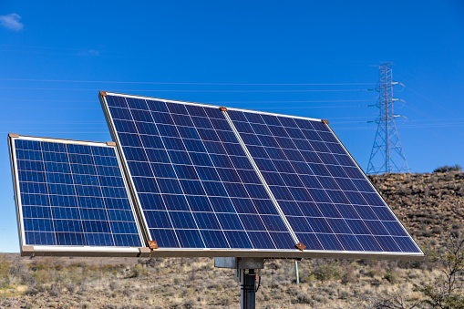 Close up on three solar panels on a make shift frame in an arid area. In the background is a high voltage electricity pylon.