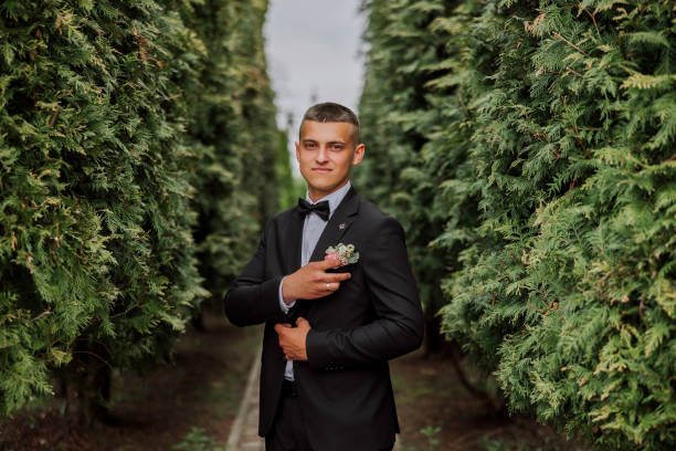 the groom in a black suit adjusts the boutonniere, poses against the background of a green tree. wedding portrait. - fashion model personal accessory suit tying ストックフォトと画像