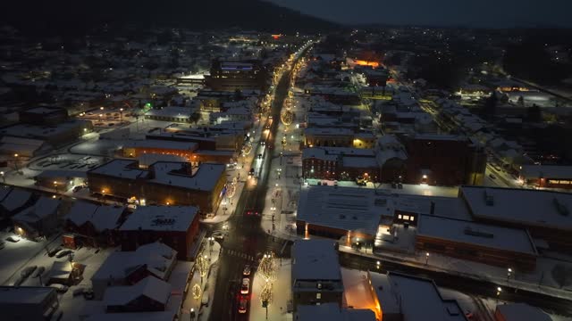 Snow covered town at night. Aerial orbit shot above lit up main street with Christmas lights on trees and quaint buildings.
