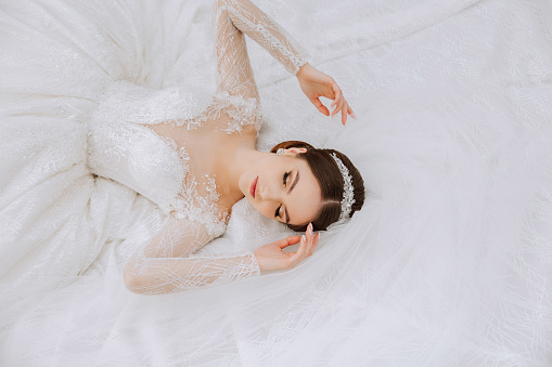 A brunette bride with a tiara in a lacy white dress with long sleeves poses against the background of her dress. On a white background. Classic hairstyle. Evening makeup. Spring wedding