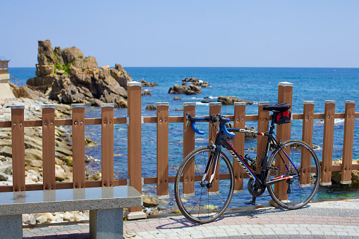 Goseong County, South Korea - July 30, 2019: A road bicycle leans against a fence on a lookout patio near Geojin 1-ri Beach and Geojin Port, offering a breathtaking view of the East Sea coast and a unique rock formation on a clear summer day.