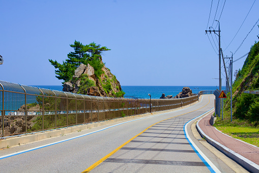 Goseong County, South Korea - July 30, 2019: A tall seaside rock towering over the coastal road and a military fence, with lush greenery atop, against a backdrop of blue sky and water, marking a serene yet protected natural landscape.