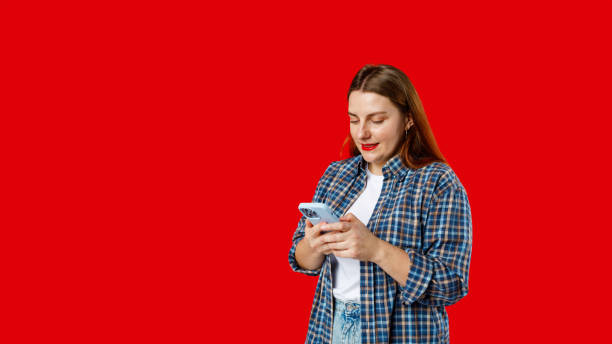 istock Smiling young brunette woman with smart phone posing isolated on red background, studio portrait. Cute beautiful young woman chatting by mobile phone 2112088083