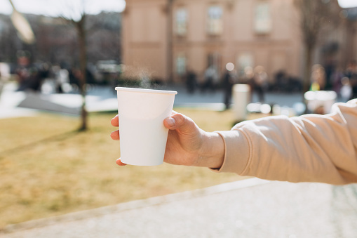 Woman hand holding white paper coffee cup outdoor. Disposable paper cup closeup. Delicious hot beverage. Blank space for text