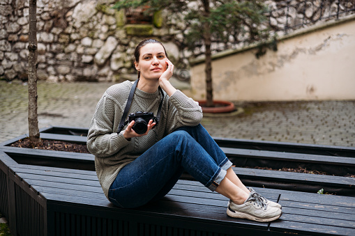 Young tourist woman smiling and using vintage camera at the city. Happy woman holding her chin and looking at the camera while sitting on bench outdoors
