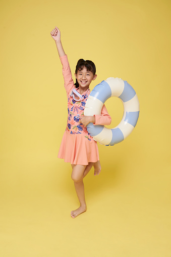 Little funny happy asian girl in swimsuit jumping up in air with inflatable ring around waist, isolated on yellow studio background. Summertime, vacation an school holidays concept