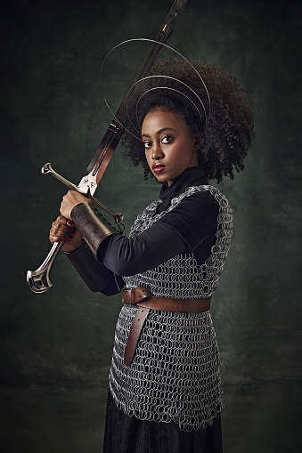 Young African woman, medieval warrior in chainmail armor with halo-like rings above head holding sword against vintage green background. Concept of history, beauty and fashion, comparison of eras