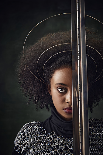 Half-face portrait of African woman with circular halos around her head and chainmail attire holding sword close face on dark vintage background. History, beauty, fashion, comparison of eras concept