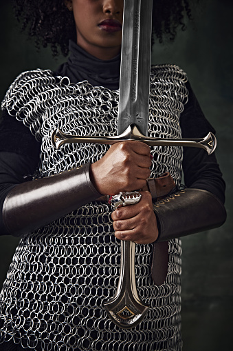 Close-up of African woman in chainmail holding sword vertically in front of face against vintage green background. Focus on hands and sword. Concept of history, beauty and fashion, comparison of eras