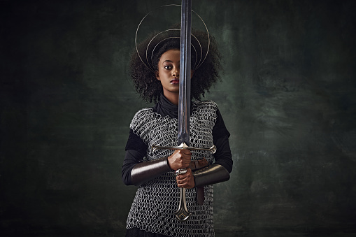 Brave young African woman, medieval warrior in chainmail armor with halo-like rings above head holding sword close to face on dark vintage background. Concept of history, emotions, comparison of eras