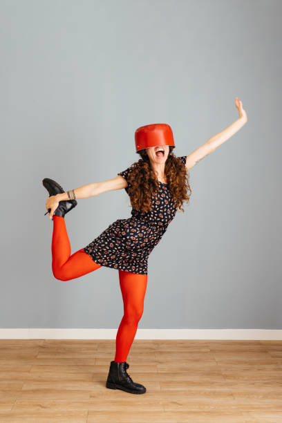 A woman dressed in a dotted dress and red tights, posing playfully with a red pot on her head, lifting her leg and arm in a whimsical fashion.
