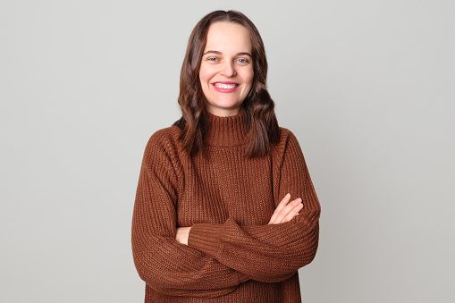 Confident satisfied Caucasian woman wearing brown sweater standing isolated over light gray background keeps her arms crossed looking at camera with toothy smile