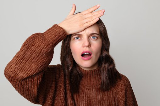 Sad despair brown haired woman wearing brown jumper standing isolated over light gray background showing facepalm gesture expressing negative emotions feeling looser