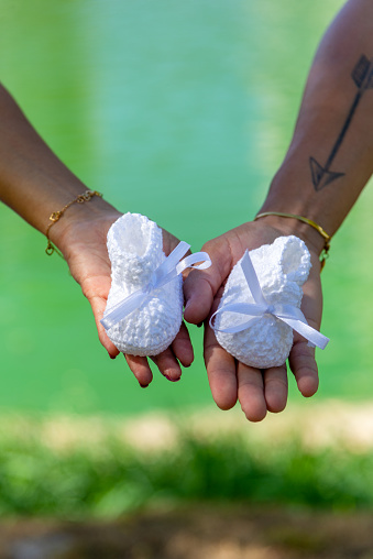Detail of a couple's hands showing their baby shoes to the camera. She is pregnant. Gender reveal. In the background a green lagoon.