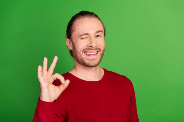 istock Photo of flirty handsome pleasant man with stubble dressed red pullover showing okey eye blinking isolated on green color background 2112256120