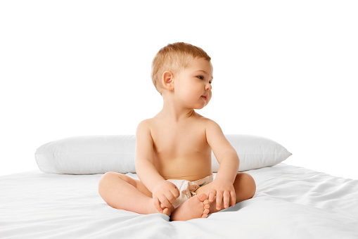 Beautiful little baby, boy, child in diaper calmly sitting on bed isolated over white studio background. Concept of childhood and family, care, parenthood, infancy and heath