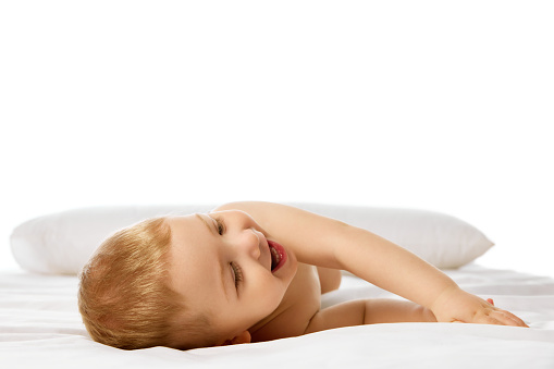 Happy child, little baby bot lying on bed on white sheets, smiling, playing isolated over white studio background. Concept of childhood and family, care, parenthood, infancy and heath