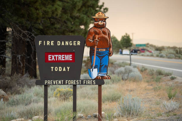 Smoky the Bear fire danger extreme Forest Service sign along highway in Sierra Nevada mountains with active fire in the distance.  Peak Fire Season.