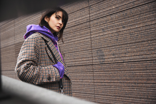 Urban photography. Beautiful young woman with short hair, wearing coat and purple hoodie, walking on empty street. Concept of street style fashion, beauty, modern trends