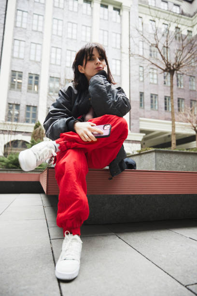 młoda kobieta w stylowej szarej kurtce i czerwonych spodniach siedzi na ławce z telefonem komórkowym na miejskim tle - seat row obrazy zdjęcia i obrazy z banku zdjęć