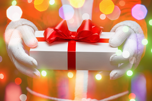 Santa Claus giving christmas present with gloved hands holding white giftbox tied up with red bow.Sparkling bubbles all around.Indoors shot,selective focus.