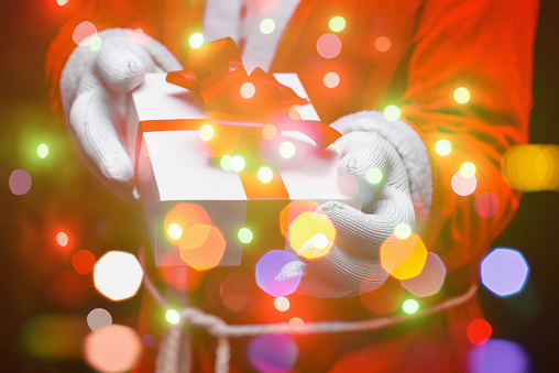 Santa Claus giving christmas present with gloved hands holding white giftbox tied up with red bow.Sparkling bubbles all around.Indoors shot,selective focus.