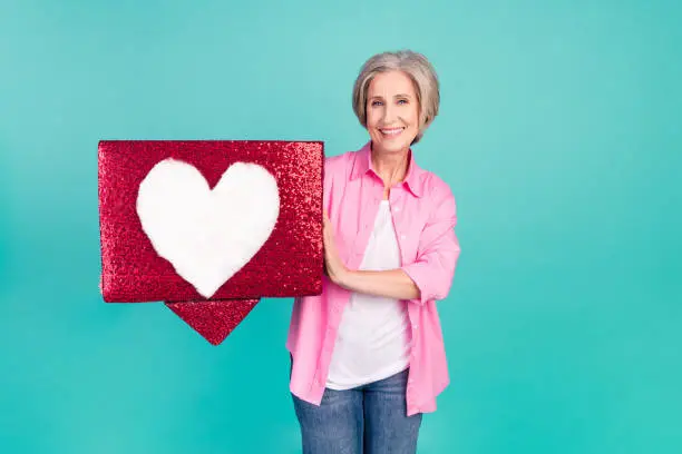 Photo of Portrait of old business woman trying to search her soulmate in dating app holding love symbol isolated on aquamarine color background