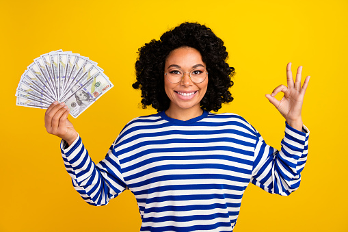Portrait of multinational good mood nice girl with chevelure in glasses hold money showing okey isolated on yellow color background.