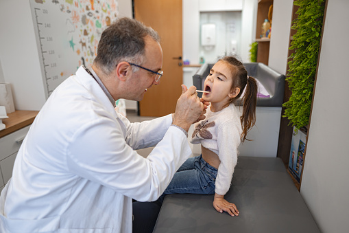 Male pediatrician examining tonsils of a little girl at the medical clinic.