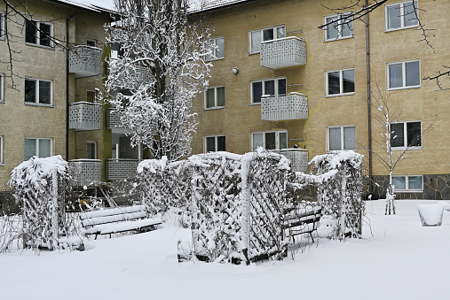 A residential area with trees and a bench covered in snow in front of a yellow apartment building with balconies