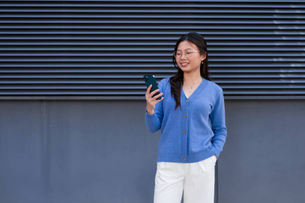 istock Young Asian woman using smart phone and standing against grey colored background 2112435080