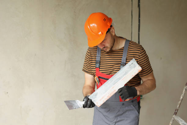 istock Worker in overalls and safety hard hat holds and prepares two spatulas for plastering concrete wall with putty using. Renovation apartment and construction new building concept. Copy place 2112457883
