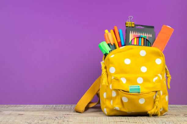 istock Yellow backpack with white polka dots with different colorful stationery on table. Purple background. Back to school. 2112458392