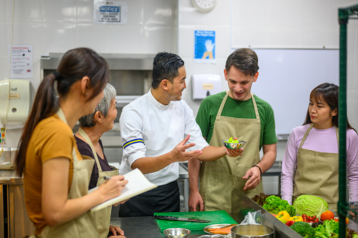 An Asian male chef giving instructions to his adult students standing around table in cooking class - Professional Cooking School