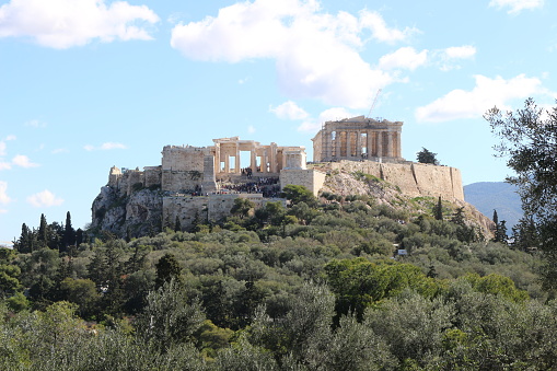 Amidst the azure expanse, the Acropolis basks in the warm embrace of summer, beckoning tourists to explore its ancient wonders amidst the picturesque backdrop of fluffy clouds