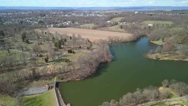 Aerial drone view of a water dam with a cemetery next to it. Mountains and sky visible in the distance.