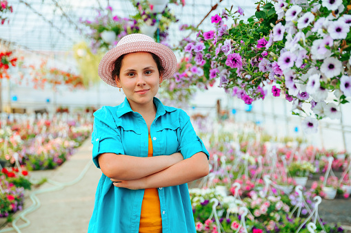 Front view of a young agronomist girl in a greenhouse, the woman crosses her arms over her chest and smiles playfully. The agronomist girl stands in a hat and shirt among the flowers.