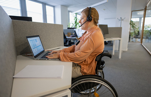 Young businesswoman in wheelchair analyzing data spreadsheet on laptop in disability accessible office. Business and career concept.